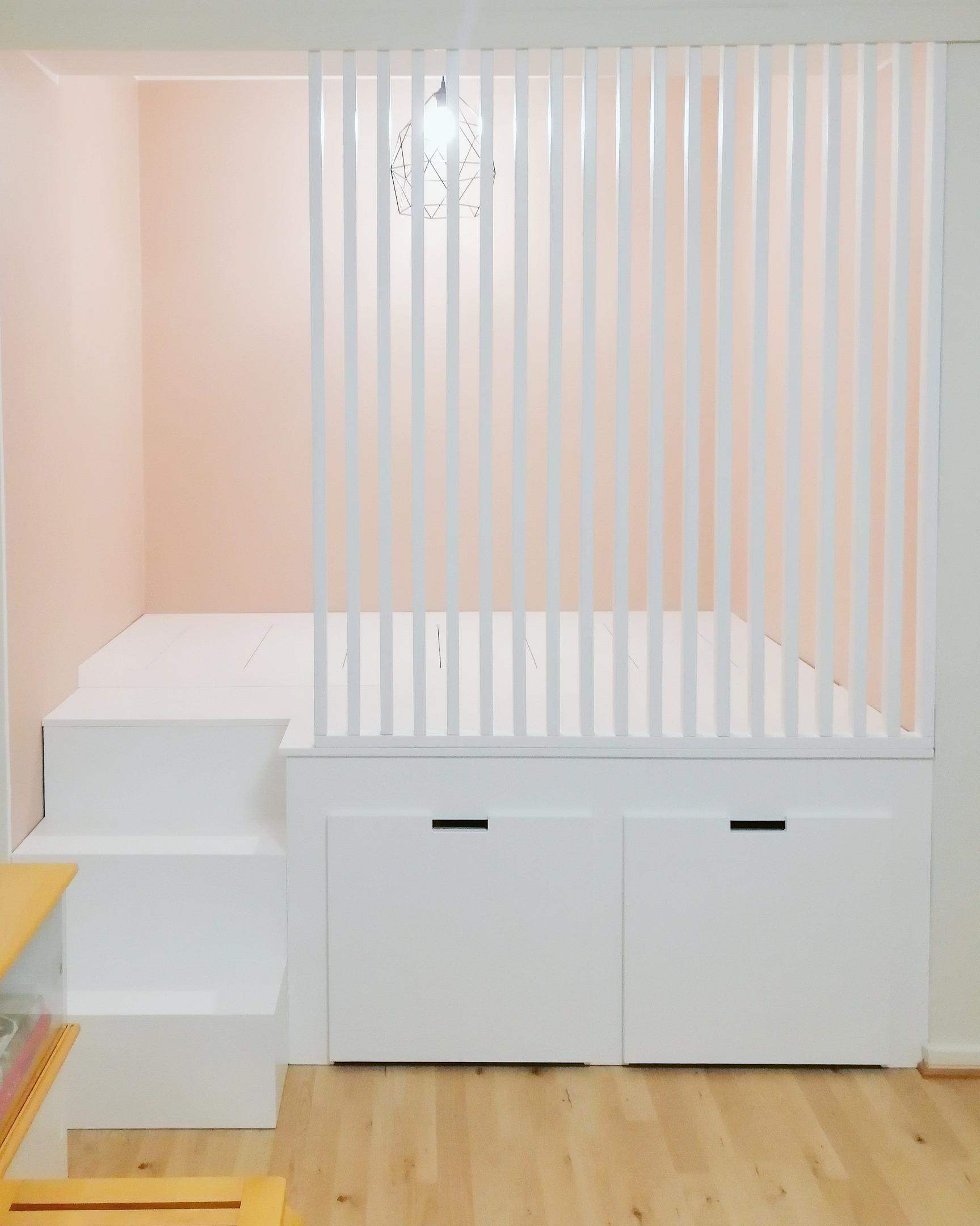 Alkovin korotettu sänky, edessä rimaseinä, portaat ja kaksi isoa pyörällistä vetolaatikkoa